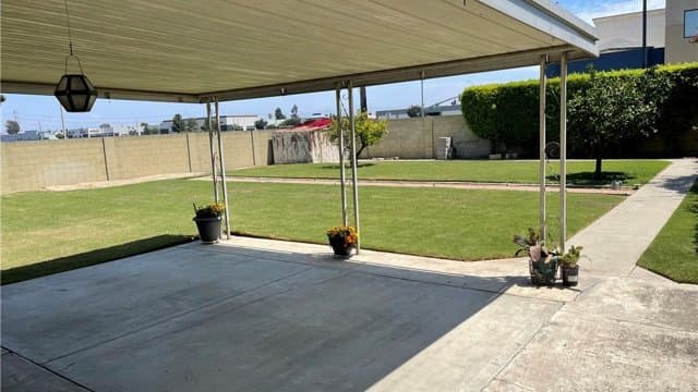 A covered concrete patio with potted plants overlooks a green lawn, a level concrete walkway, and a block wall in the backyard.