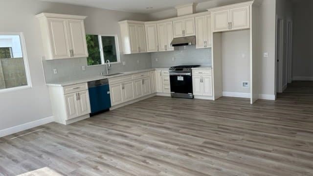 A modern, empty kitchen and adjacent open living space featuring light cream cabinets, white countertops, light gray subway tile backsplash, stainless steel appliances, and light wood-look laminate flooring.