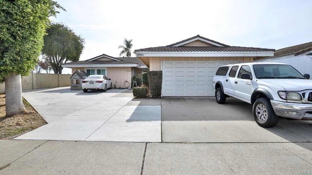 A single-story, light-colored house with a two-car garage and a tile roof, featuring a white sedan and a white pickup truck parked in the concrete driveway, flanked by a large tree on the left.