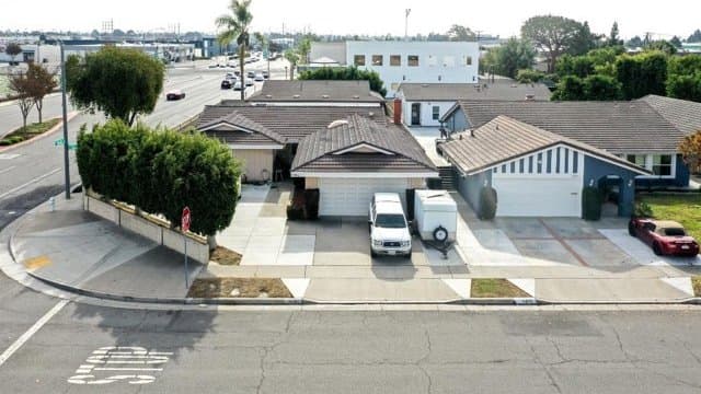Aerial view of single-story residential homes along a street corner, with cars parked in driveways. Commercial buildings and distant structures are visible in the background.
